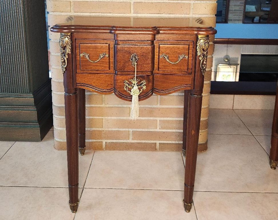 Mesita de noche antigua de madera con cajones, detalles de latón y patas delgadas contra una pared de ladrillos.