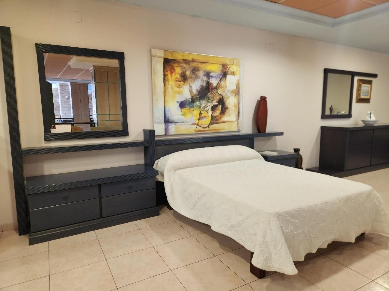 Conjunto de muebles de dormitorio: cama con ropa de cama blanca, mesita de noche, cómoda, espejos y cuadro abstracto.