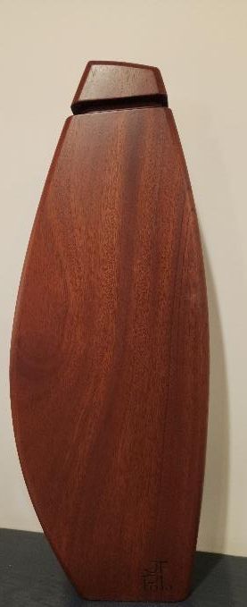 Un jarrón de madera alto, con forma de lágrima y tapa cuadrada, reposa sobre una superficie oscura contra una pared neutra.