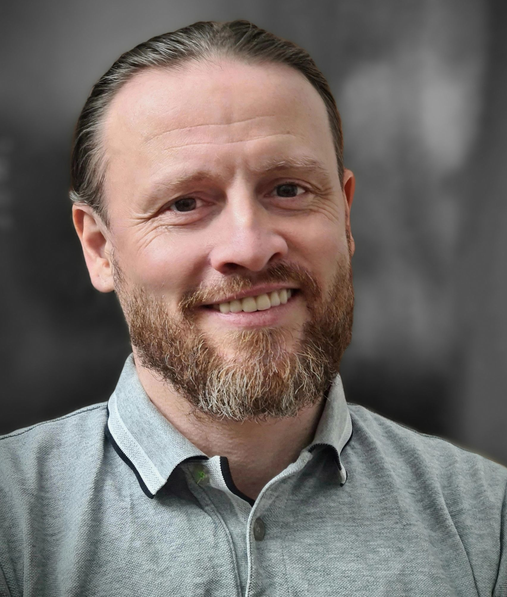 Mann mit hellbraunem Bart und Haaren, lächelnd, trägt ein graues Hemd, vor verschwommenem Hintergrund.