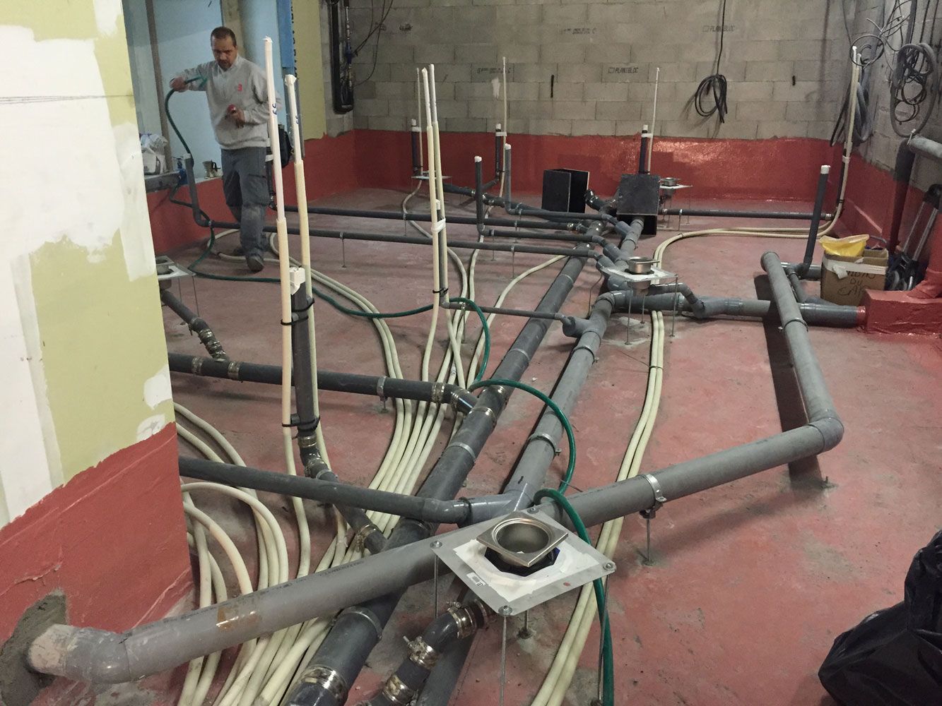 Un homme inspecte un système de plomberie complexe sur un chantier de construction ; des tuyaux et des câbles sont éparpillés sur le sol.