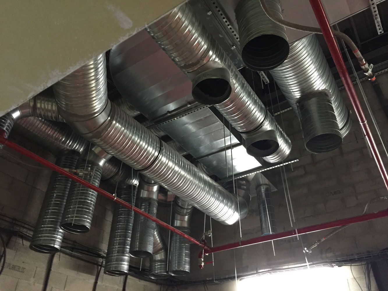 Conduits de ventilation en métal galvanisé au plafond, avec tuyaux d'extincteurs automatiques rouges, dans un bâtiment inachevé.