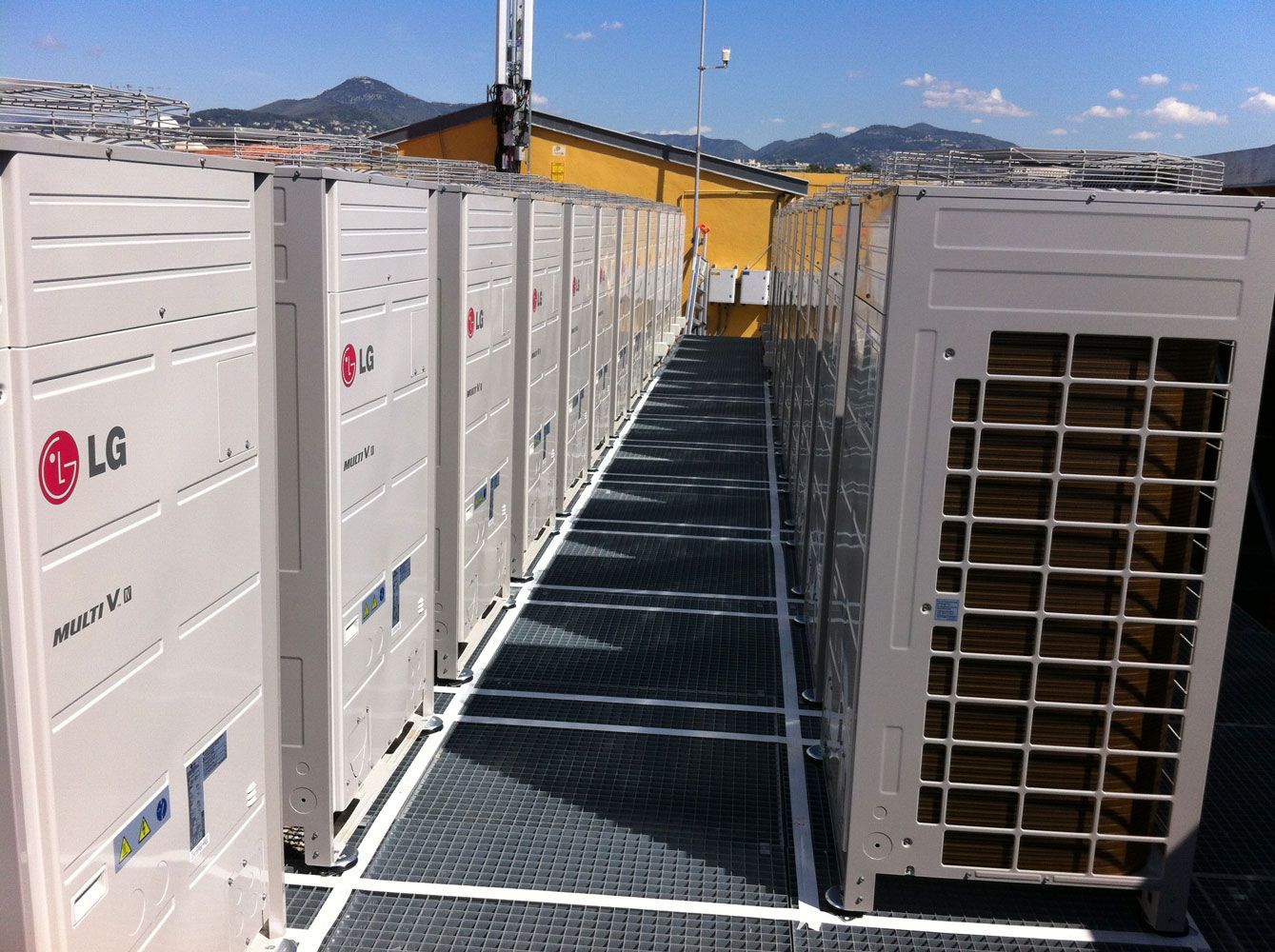 Rangée d'unités de climatisation LG sur un toit-terrasse avec une passerelle métallique, surplombant une ville et des montagnes.