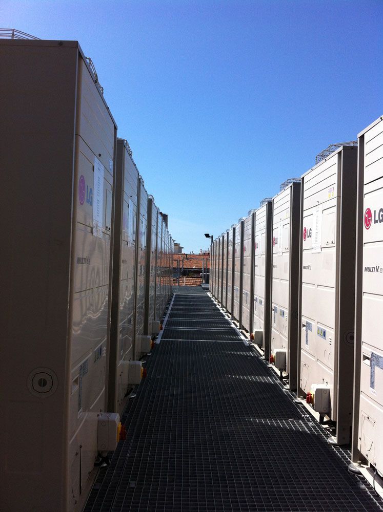 Une rangée de grands climatiseurs extérieurs sur une passerelle métallique, sous un ciel bleu azur.