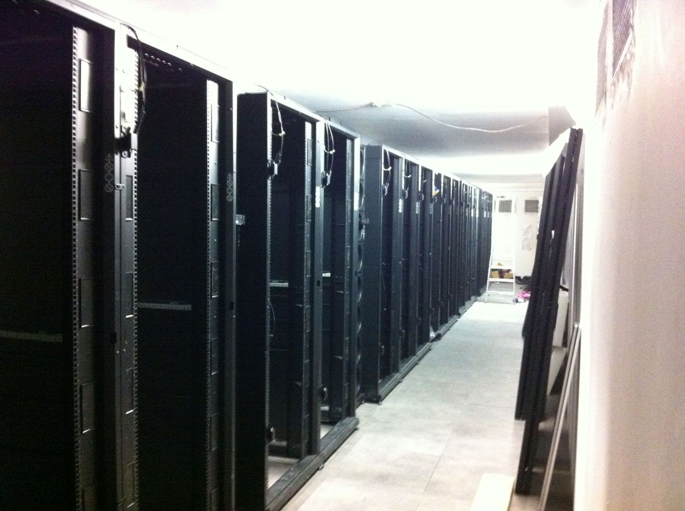 Rangées de baies de serveurs noires vides dans un centre de données, éclairées par un éclairage zénithal. Sol en béton.