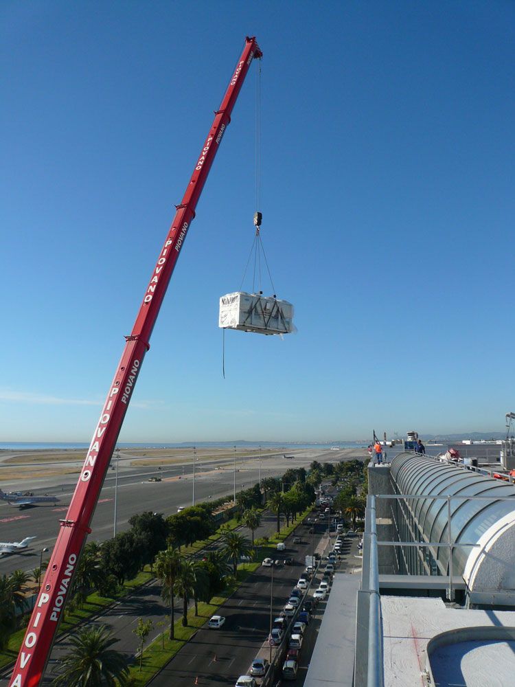 Une grue rouge soulève un conteneur blanc au-dessus d'un bâtiment, d'un aéroport et d'un ciel bleu en arrière-plan.