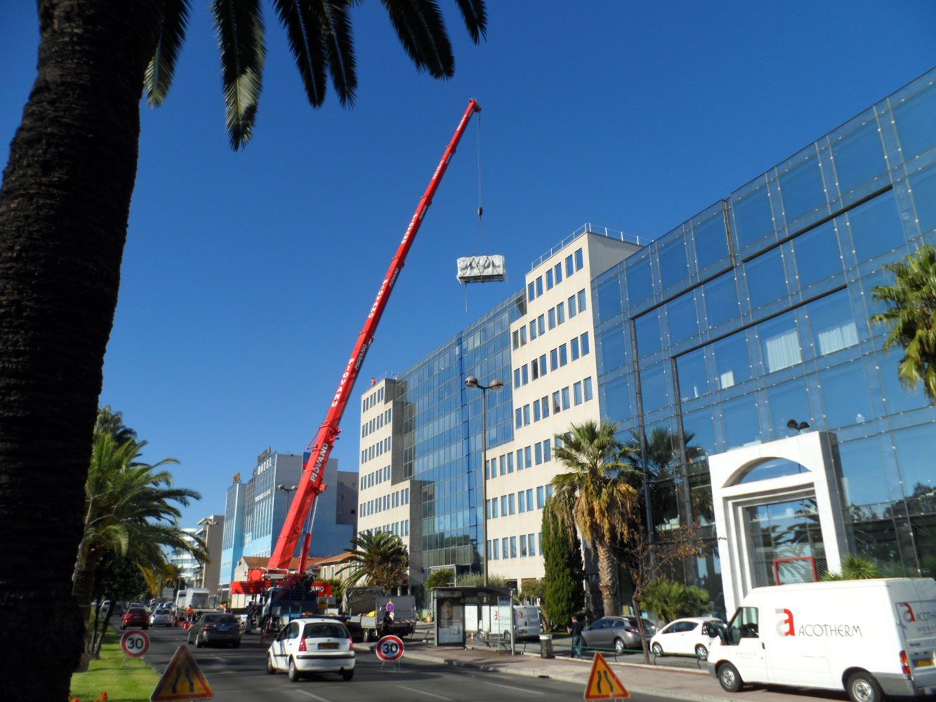 Une grue rouge soulève un objet près d'un immeuble aux façades de verre et de palmiers dans une rue ensoleillée.