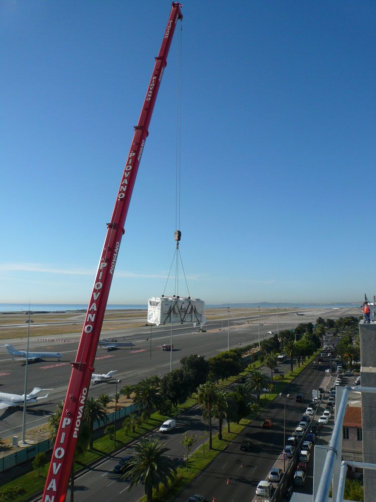 Une grue rouge soulève une structure blanche, le long d'une route côtière et d'une piste d'aéroport, sous un ciel bleu.