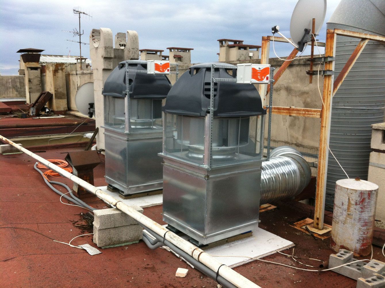 Deux unités de climatisation métalliques sur un toit rouge, entourées d'autres équipements et de tuyaux. Ciel nuageux.