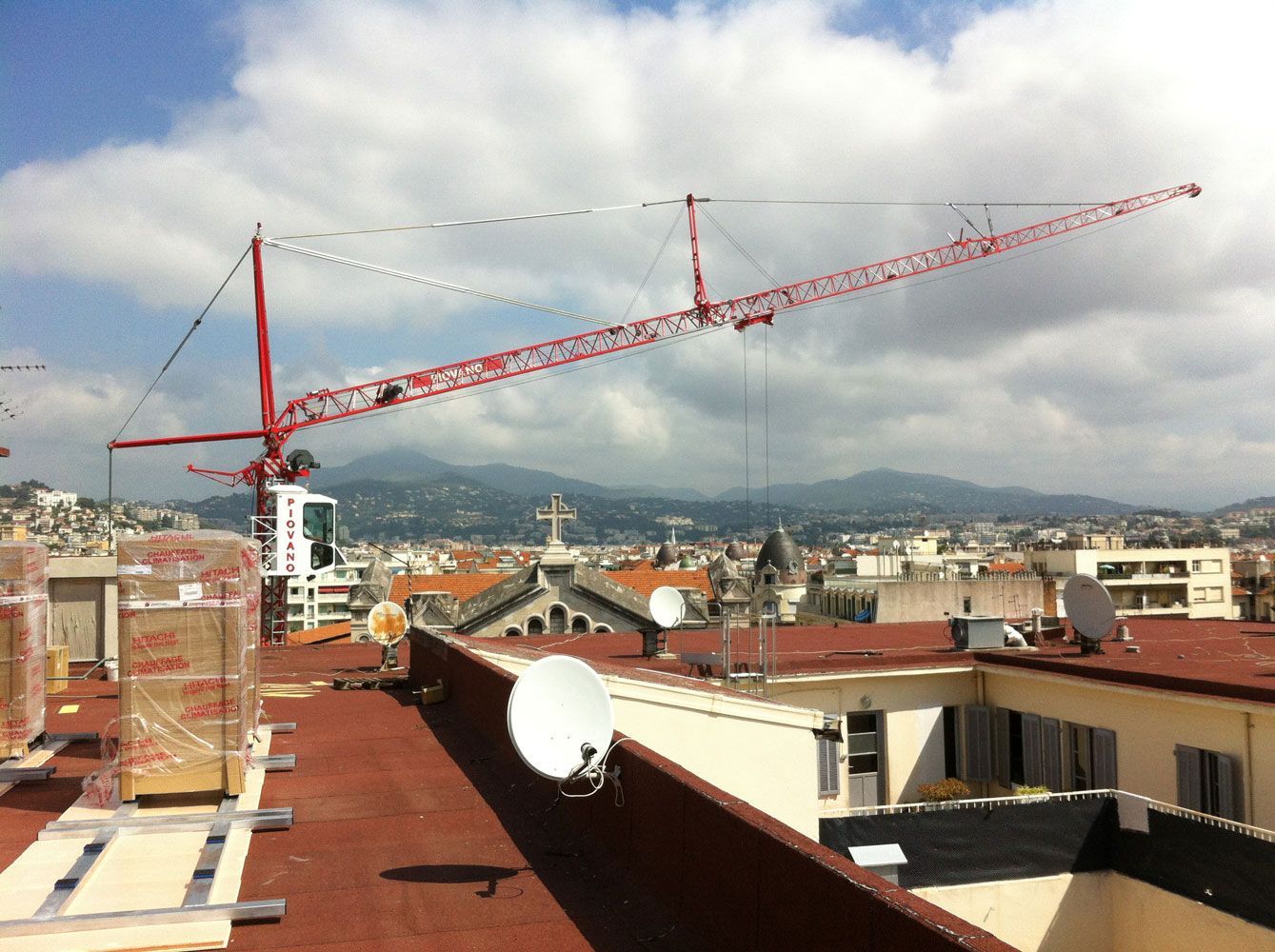 Grue de chantier rouge dominant l'horizon urbain, avec ses immeubles et ses montagnes, par une journée partiellement nuageuse.