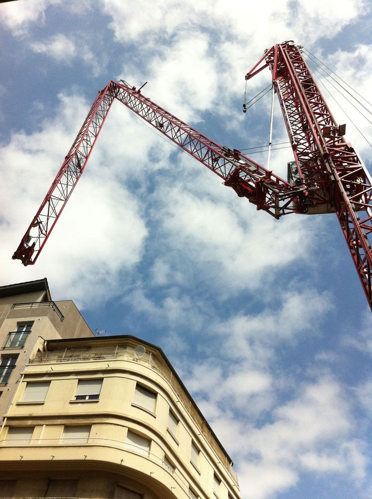 Grue de chantier rouge surplombant un bâtiment, se détachant sur un ciel bleu nuageux.