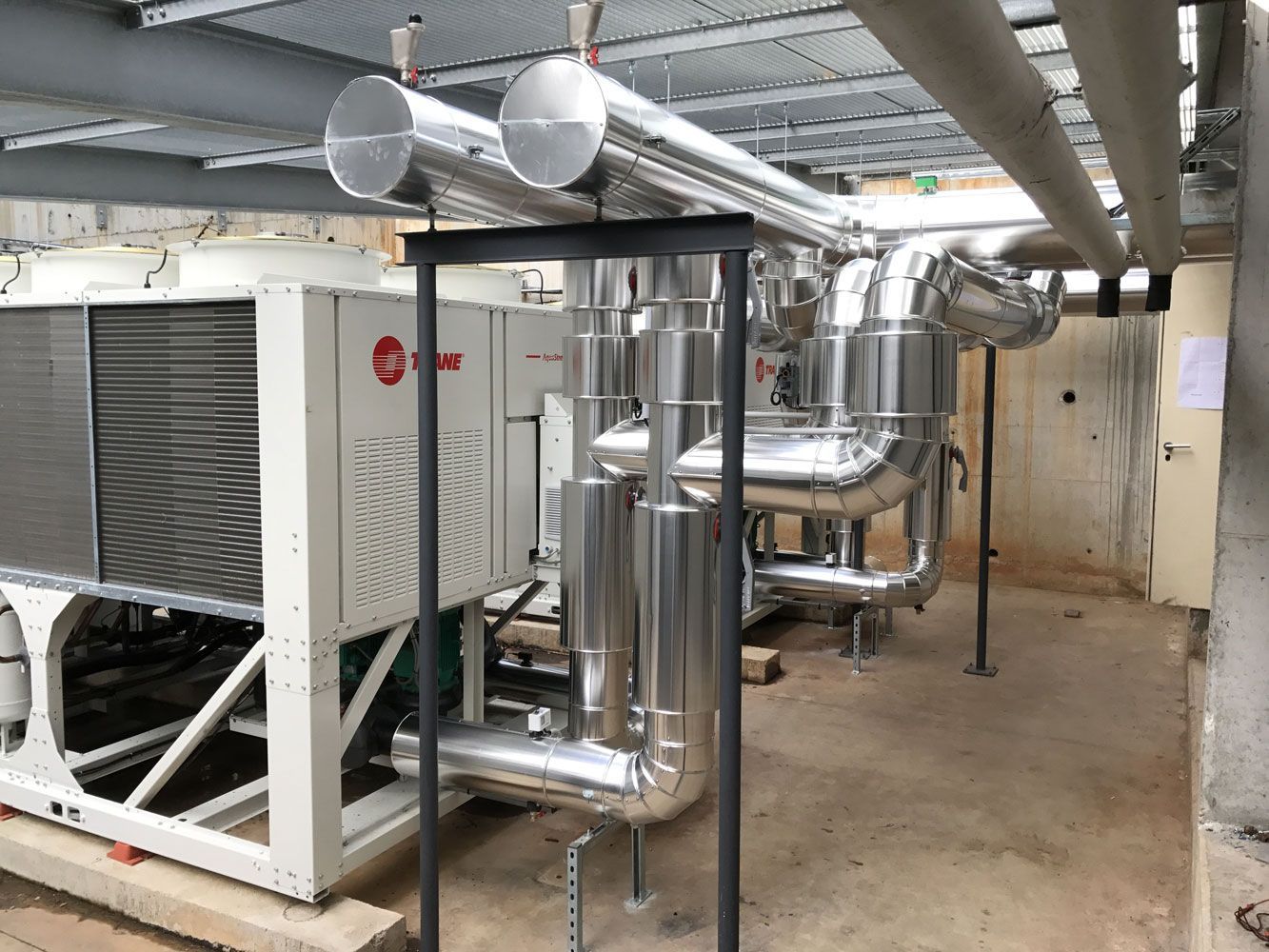 Unité de climatisation industrielle et tuyaux isolés dans une pièce en béton.