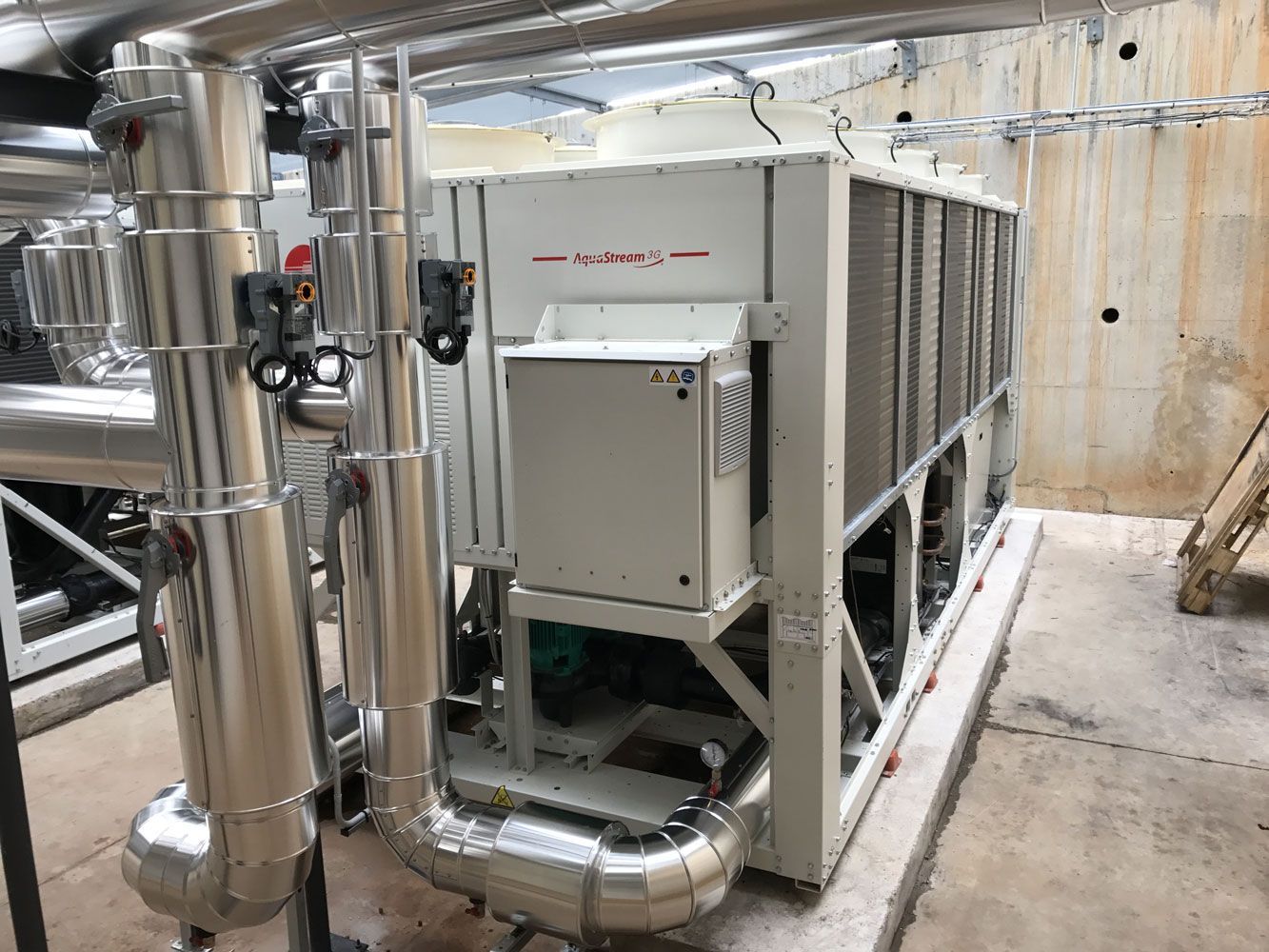 Système CVC industriel avec tuyaux argentés et grande machine blanche dans un espace en béton.