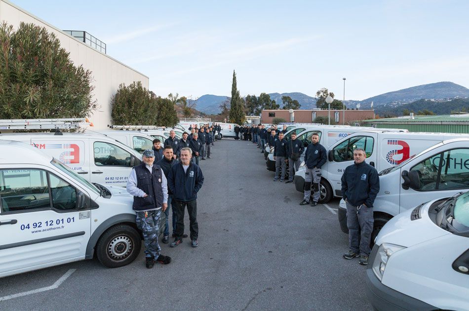 Un groupe d'ouvriers en uniforme se tient à côté d'une flotte de fourgonnettes blanches, à l'extérieur, devant un bâtiment.