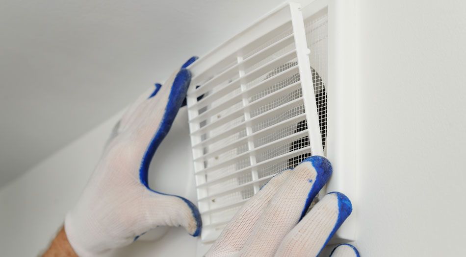 Des mains gantées de travail retirent une grille d'aération blanche d'un mur.