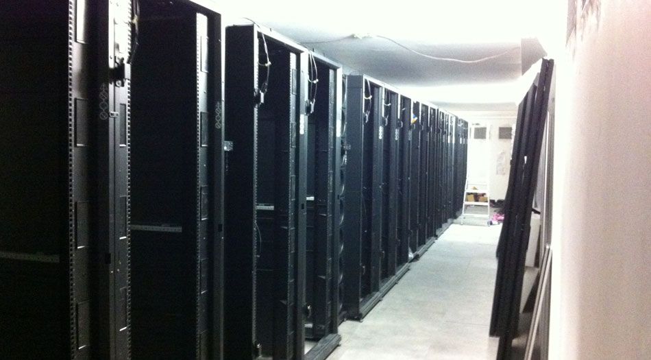 Des baies de serveurs noires et vides dans un long couloir, éclairées par des plafonniers. Murs et sol blancs.