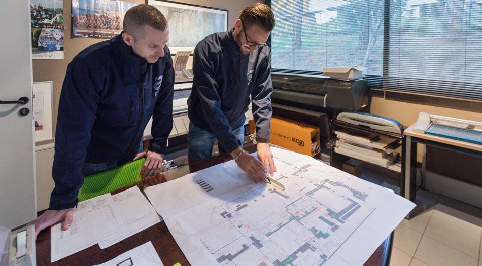 Deux hommes dans une pièce, examinant des plans sur une table. L'un désigne les plans du doigt tandis que l'autre observe.