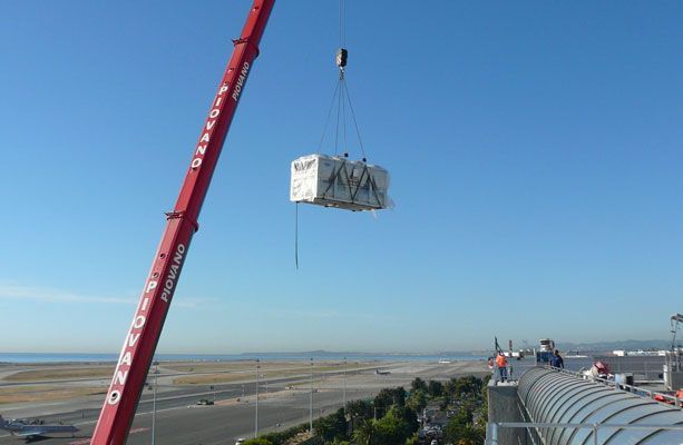 Une grue soulève un grand caisson blanc au-dessus d'un bâtiment où l'on aperçoit des ouvriers, un ciel bleu et un paysage côtier.