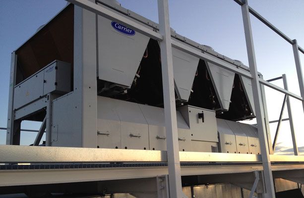 Un grand groupe frigorifique Carrier blanc à panneaux inclinés est installé sur un toit doté d'une rambarde métallique, avec un ciel bleu en arrière-plan.