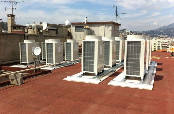 Unités de climatisation sur un toit, blanches et grises sur fond de ciel bleu, paysage urbain en arrière-plan.