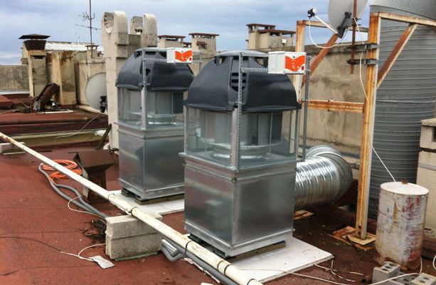 Deux grandes unités de ventilation métalliques sur un toit. Une gaine et d'autres équipements de toiture sont présents. Ciel nuageux.