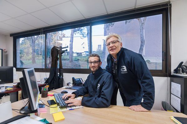 Deux hommes dans un bureau lumineux ; l'un tapant sur un ordinateur, l'autre debout à côté de lui, tous deux souriants.