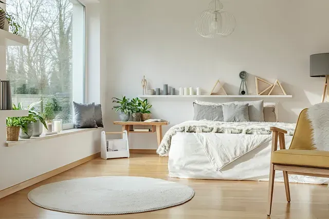 Acogedor dormitorio con ropa de cama blanca, una alfombra redonda y plantas cerca de una gran ventana.
