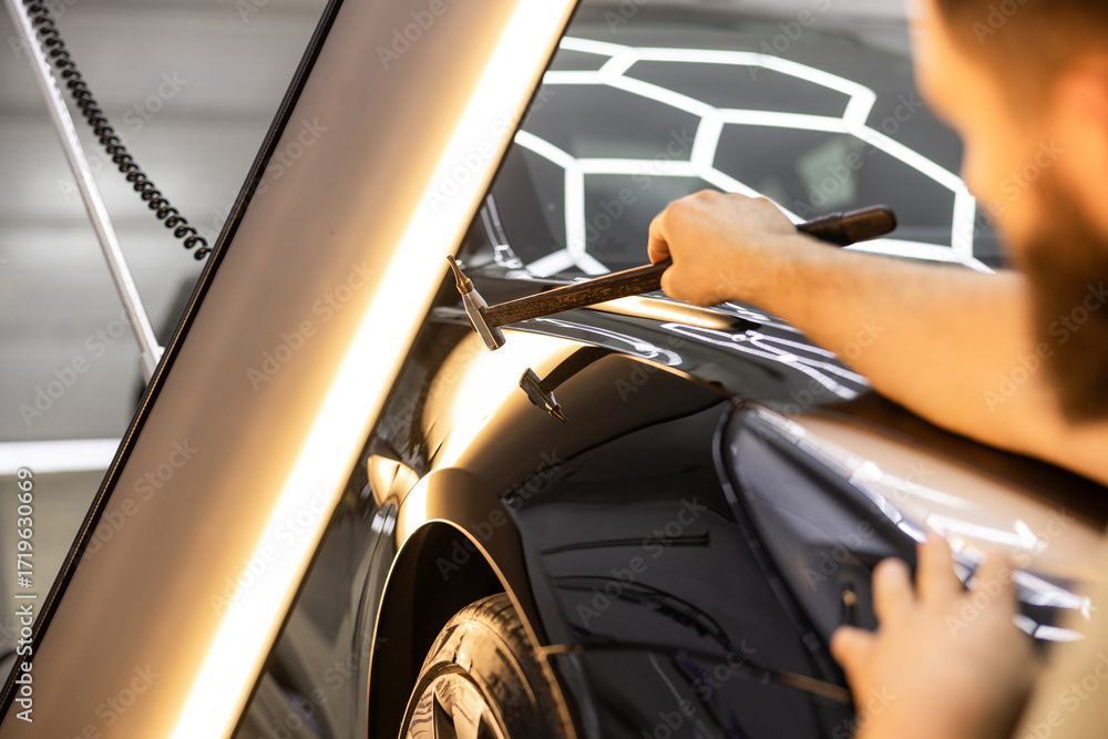 Hände bearbeiten ein Werkzeug an einer schwarzen Autokarosserie in einer Werkstatt, helles Licht und ein sechseckiger Lichthintergrund.