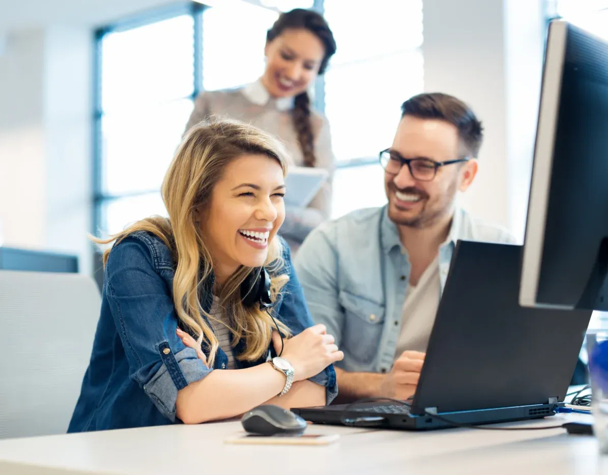 Zwei Kollegen lächeln in einem hellen Büro auf einen Laptop, im Hintergrund ist eine dritte Person verschwommen zu erkennen.