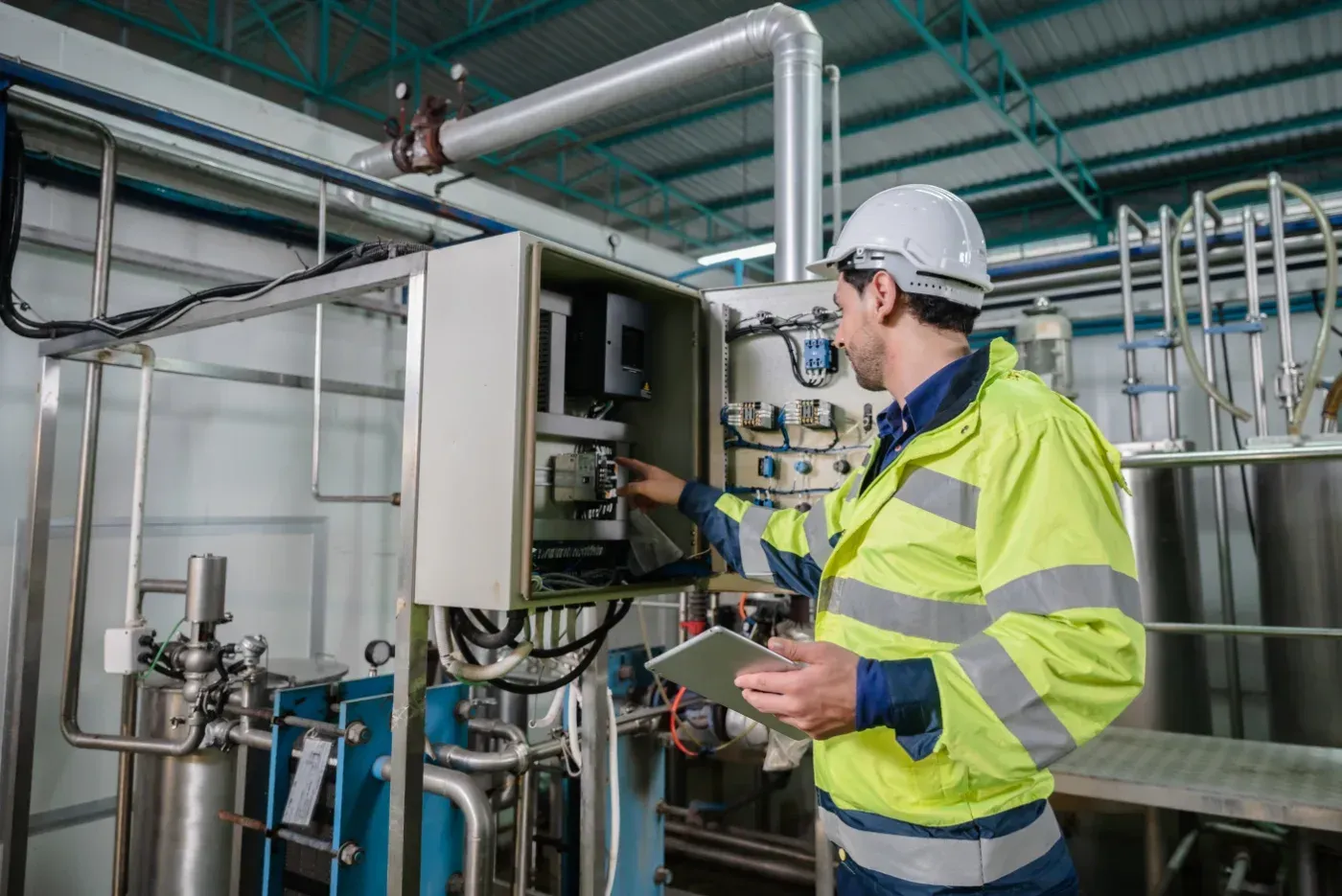 Techniker in gelber Sicherheitsjacke inspiziert industrielles Bedienfeld in einer Fabrik mit Metallrohrleitungen