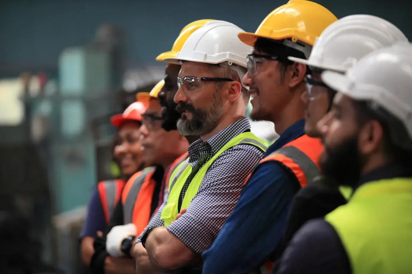 Arbeiter mit Schutzhelmen und Sicherheitswesten stehen in einer Fabrik und blicken aufmerksam nach vorn.