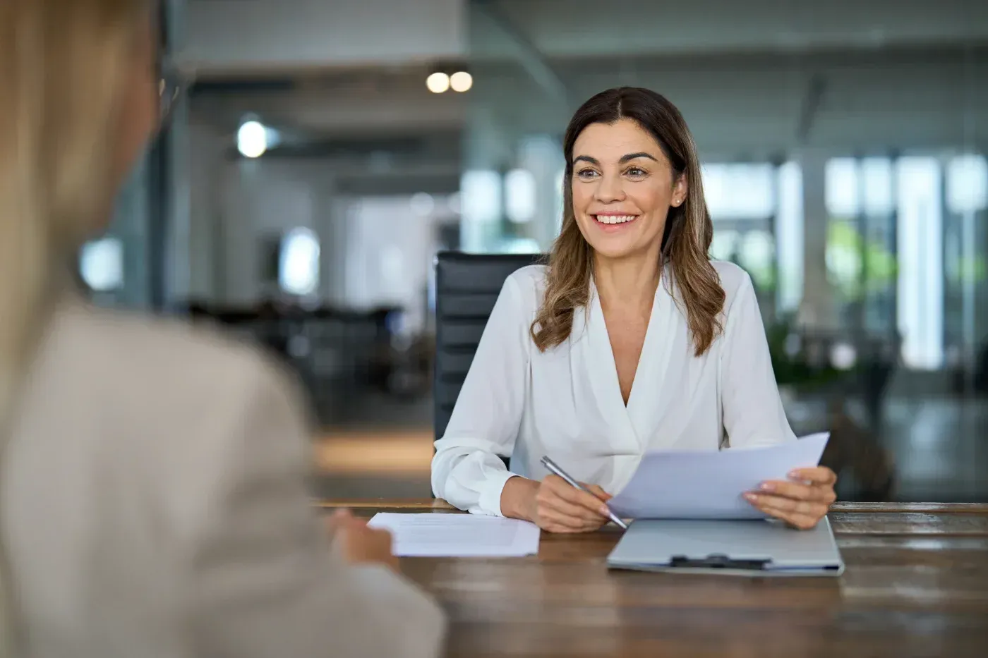 Eine Frau in weißer Bluse lächelt und liest in einem modernen Büro an einem Schreibtisch gegenüber von Papieren.