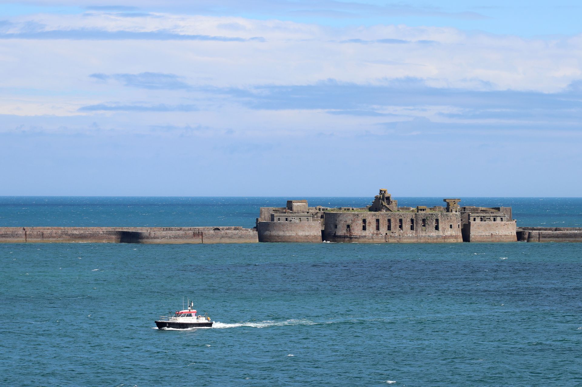 Petit bateau sur la mer bleue près d'un fort en pierre sous un ciel nuageux.