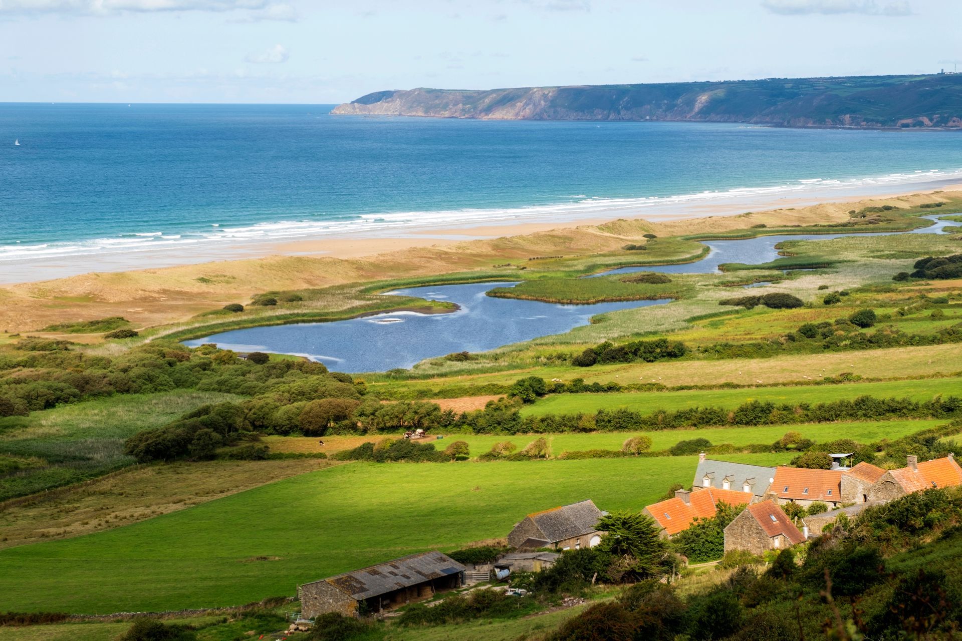 Paysage côtier avec des champs verdoyants, des étangs, une plage de sable et un océan bleu sous un ciel dégagé.