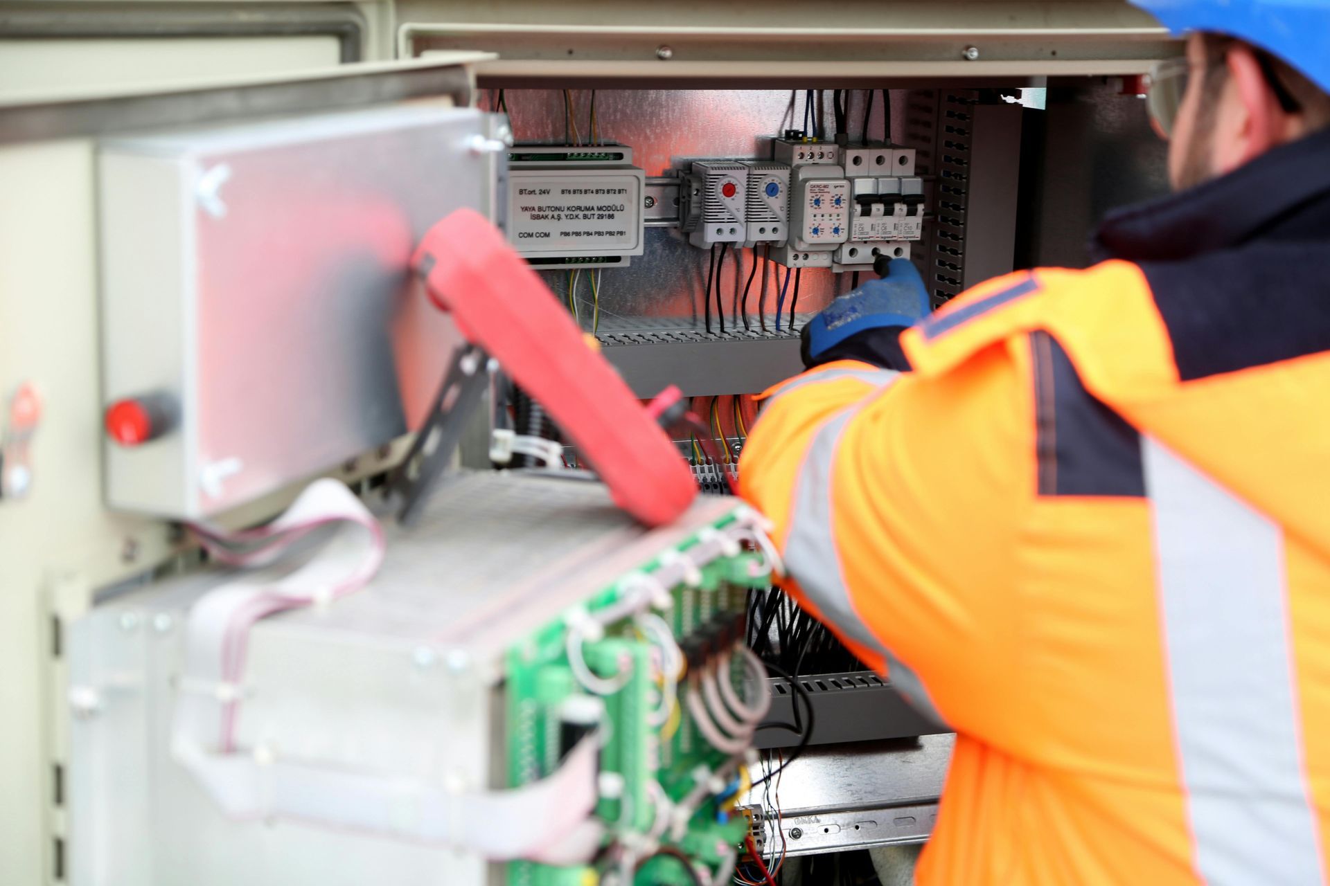 Technicien en veste de sécurité orange travaillant à l'intérieur d'un tableau électrique avec câblage et disjoncteurs.