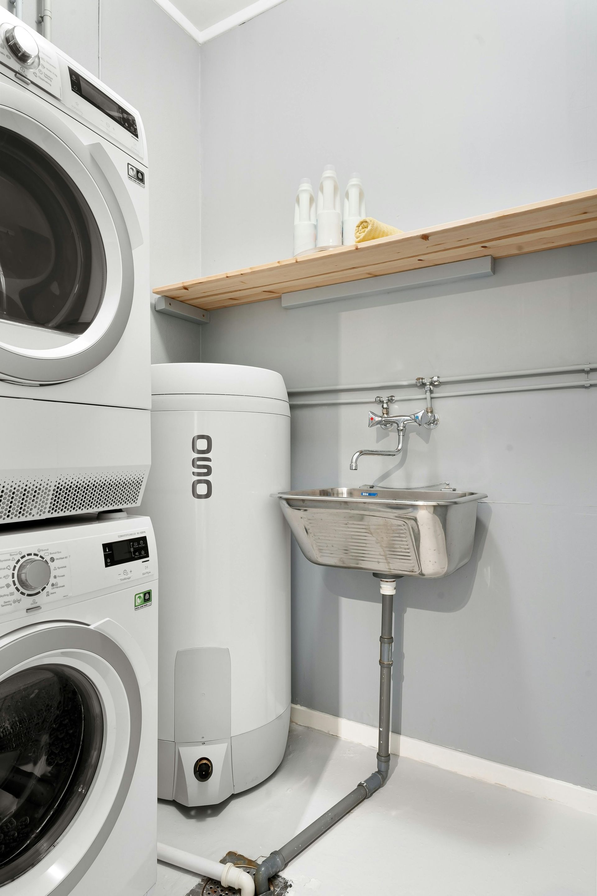 Buanderie avec lave-linge et sèche-linge superposés, évier, chauffe-eau et étagère murale en bois.