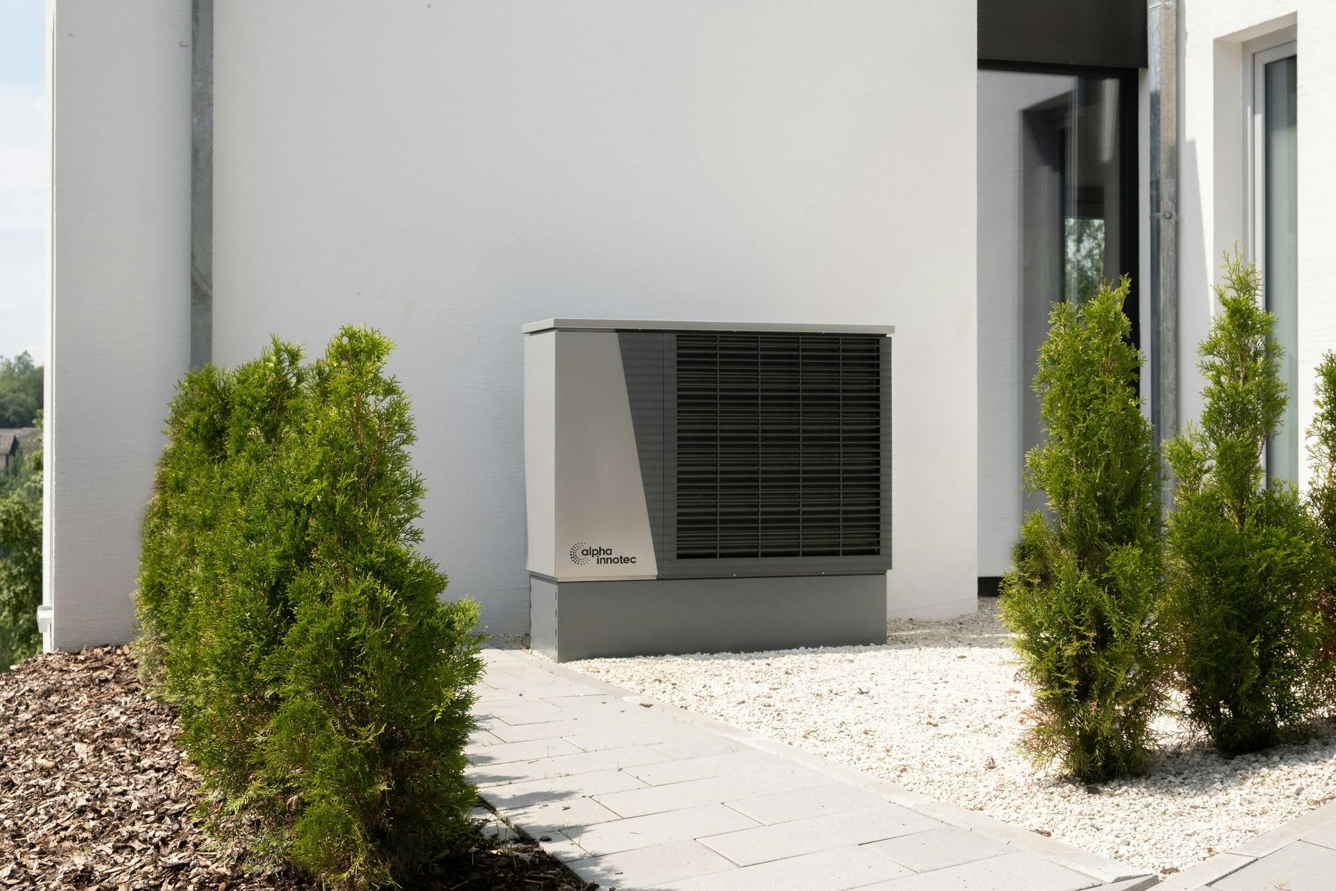 Unité de climatisation extérieure à côté d'un bâtiment blanc, encadrée par de petits arbustes verts et un chemin de gravier.