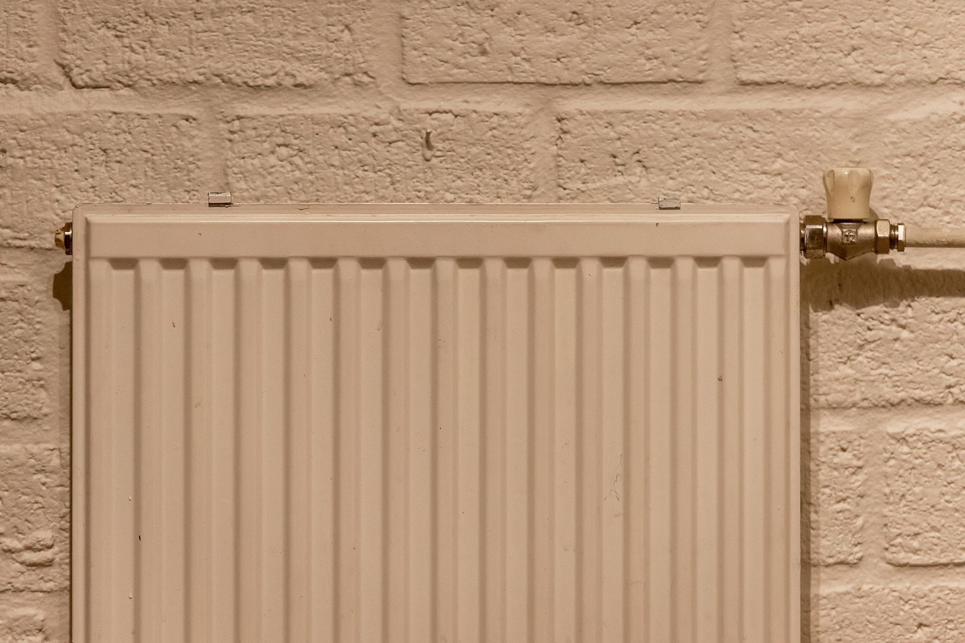 Radiateur beige fixé sur un mur en briques claires près d'une vanne de tuyauterie.