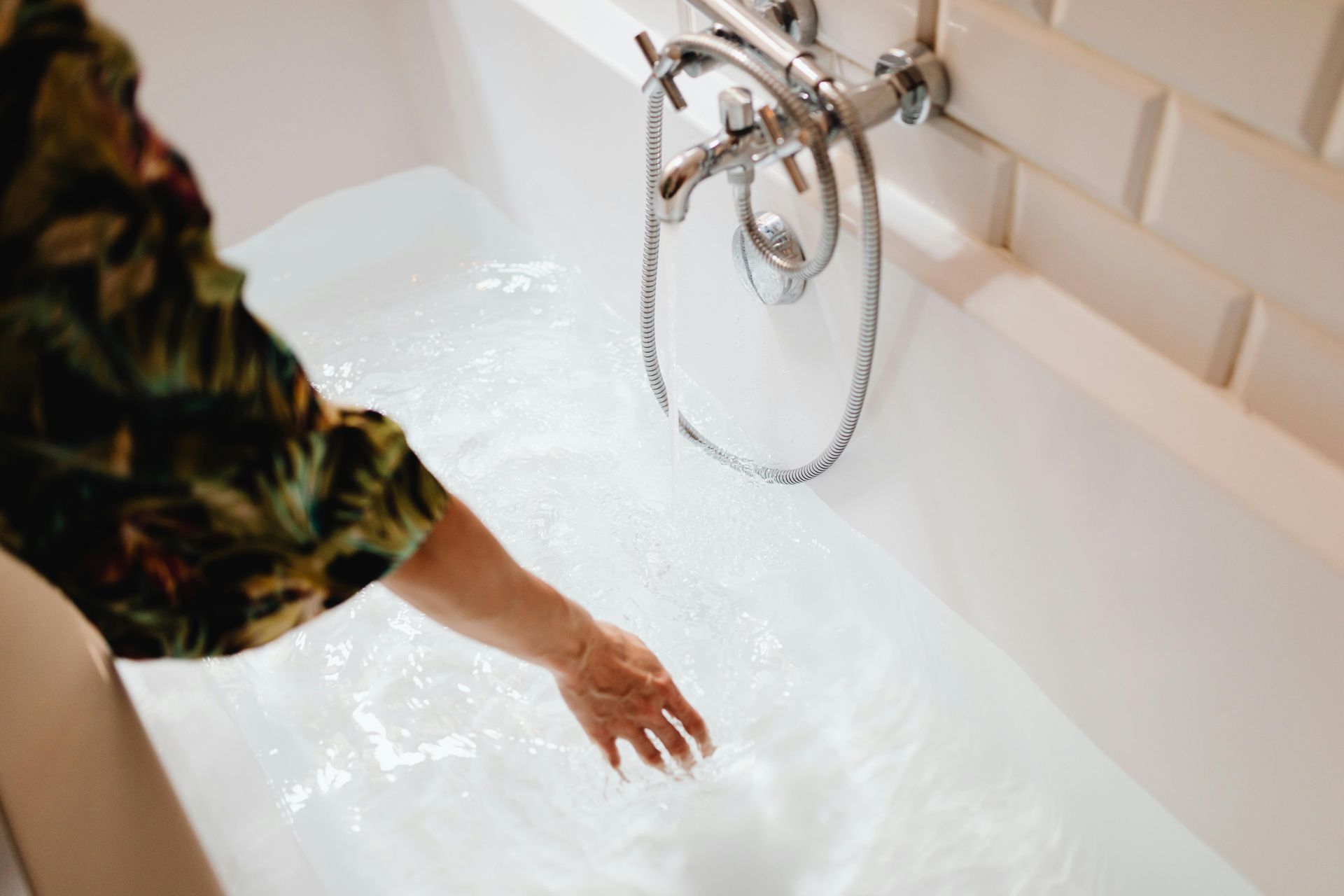 Une main plonge dans une baignoire blanche sous un robinet chromé d'eau courante.