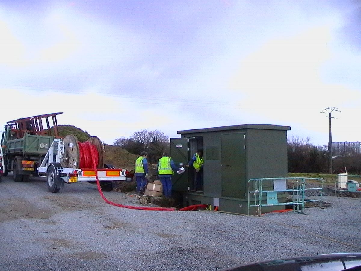 Des ouvriers installent des câbles dans un boîtier électrique, reliés à un camion transportant des bobines de câble. Journée nuageuse.