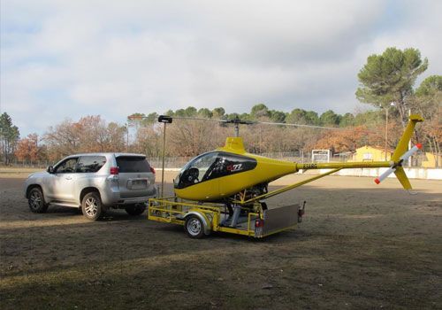 Un hélicoptère jaune sur une remorque, tracté par un SUV argenté, sur un champ herbeux.