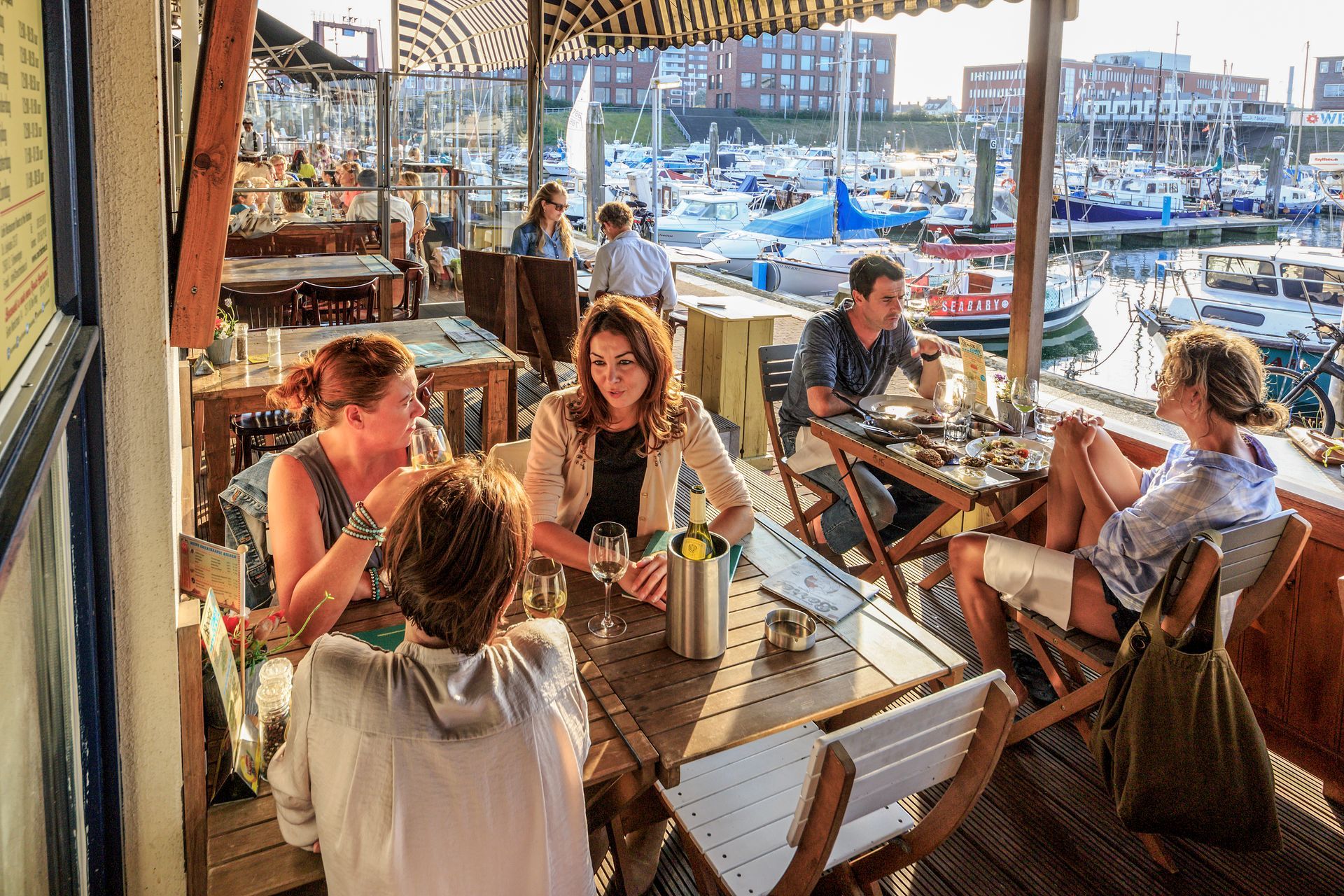 Mensen die dineren in een restaurant aan het water, boten op de achtergrond, zonnige dag.