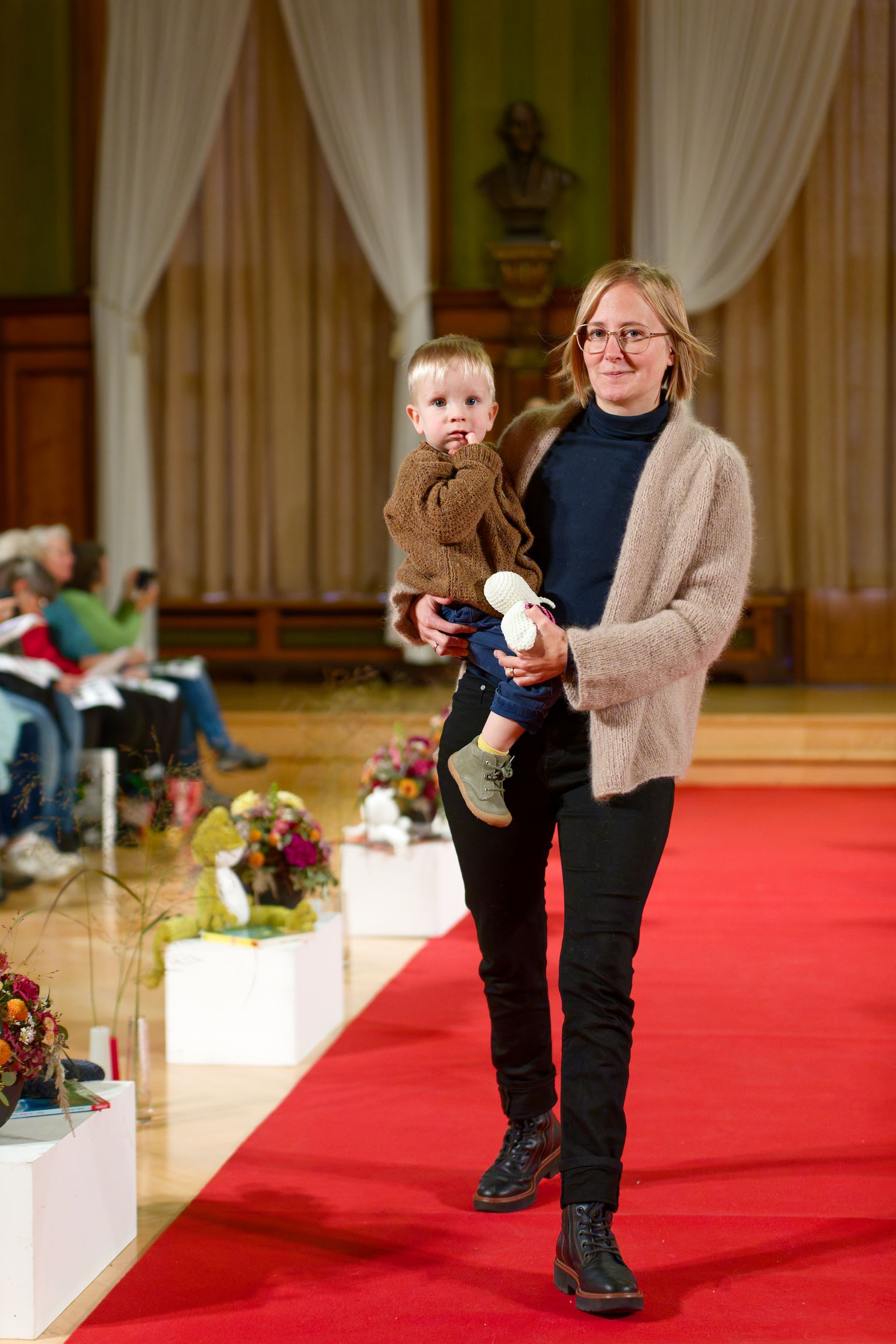 Eine Frau mit einem Kind auf dem Arm schreitet über einen roten Teppich; es handelt sich um eine Modenschau.