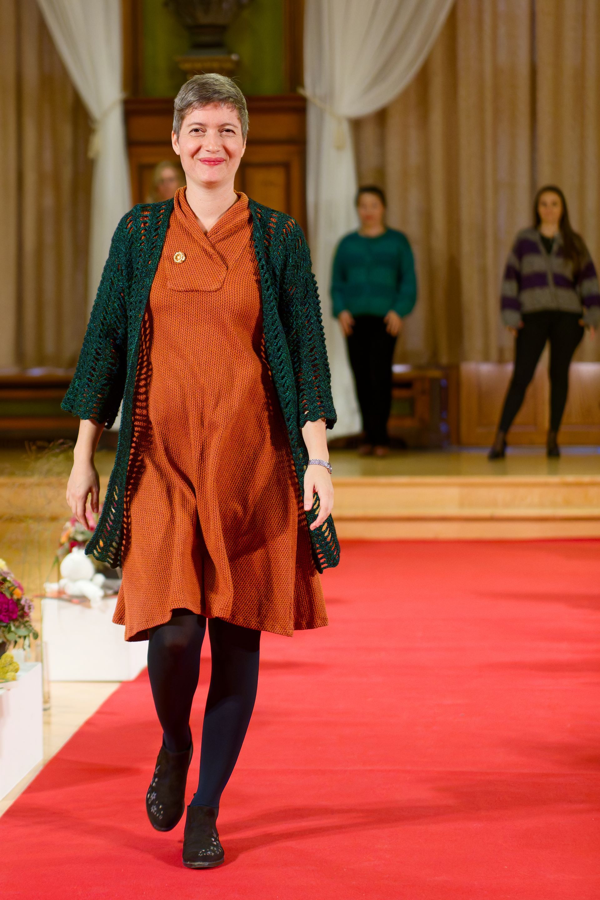Eine Frau in einem rostfarbenen Kleid und einer grünen Strickjacke schreitet lächelnd über einen roten Teppich.