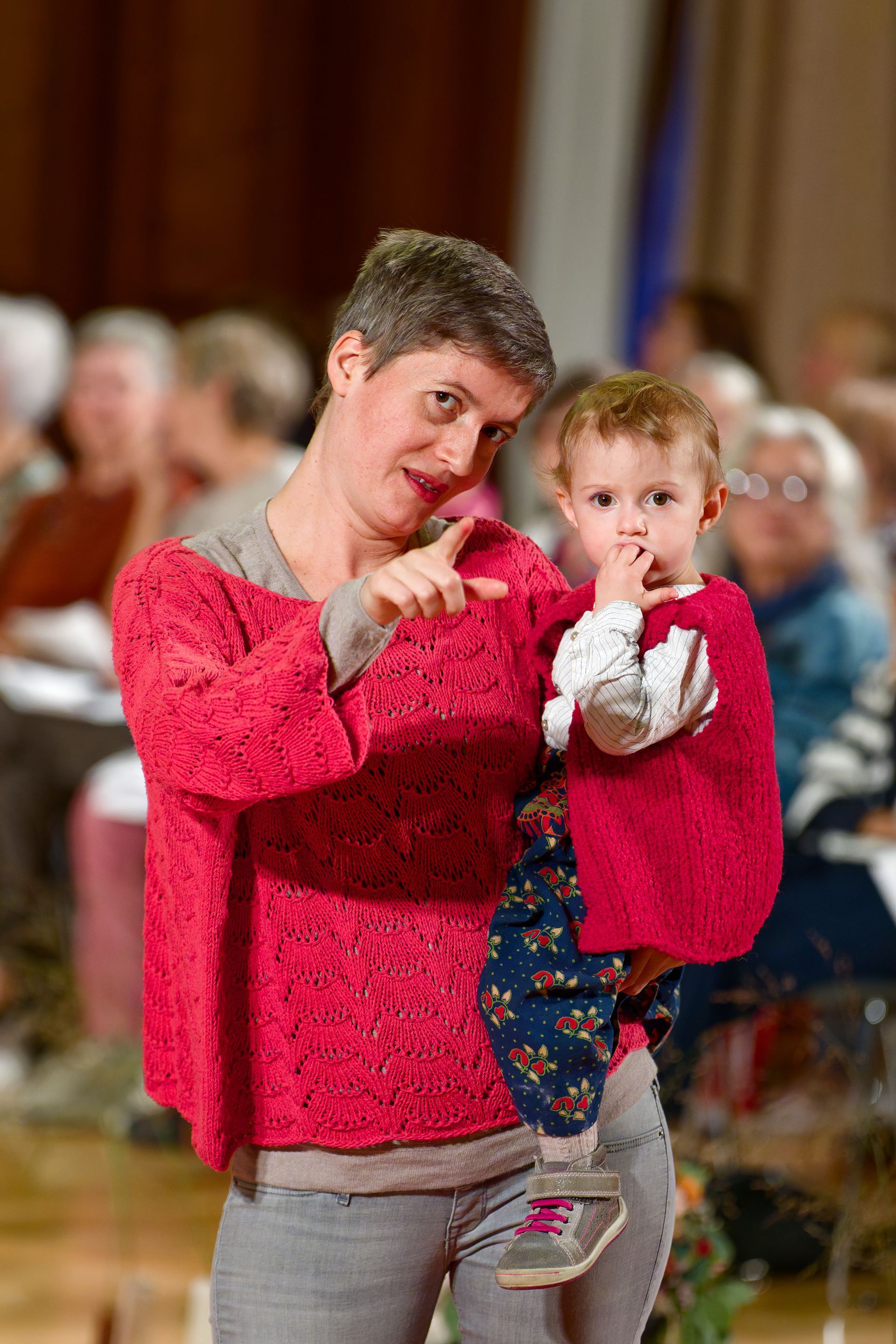 Frau in rotem Pullover zeigt mit dem Finger, hält ein Kind in Rot im Arm, in einem Raum mit anderen Personen im Hintergrund.