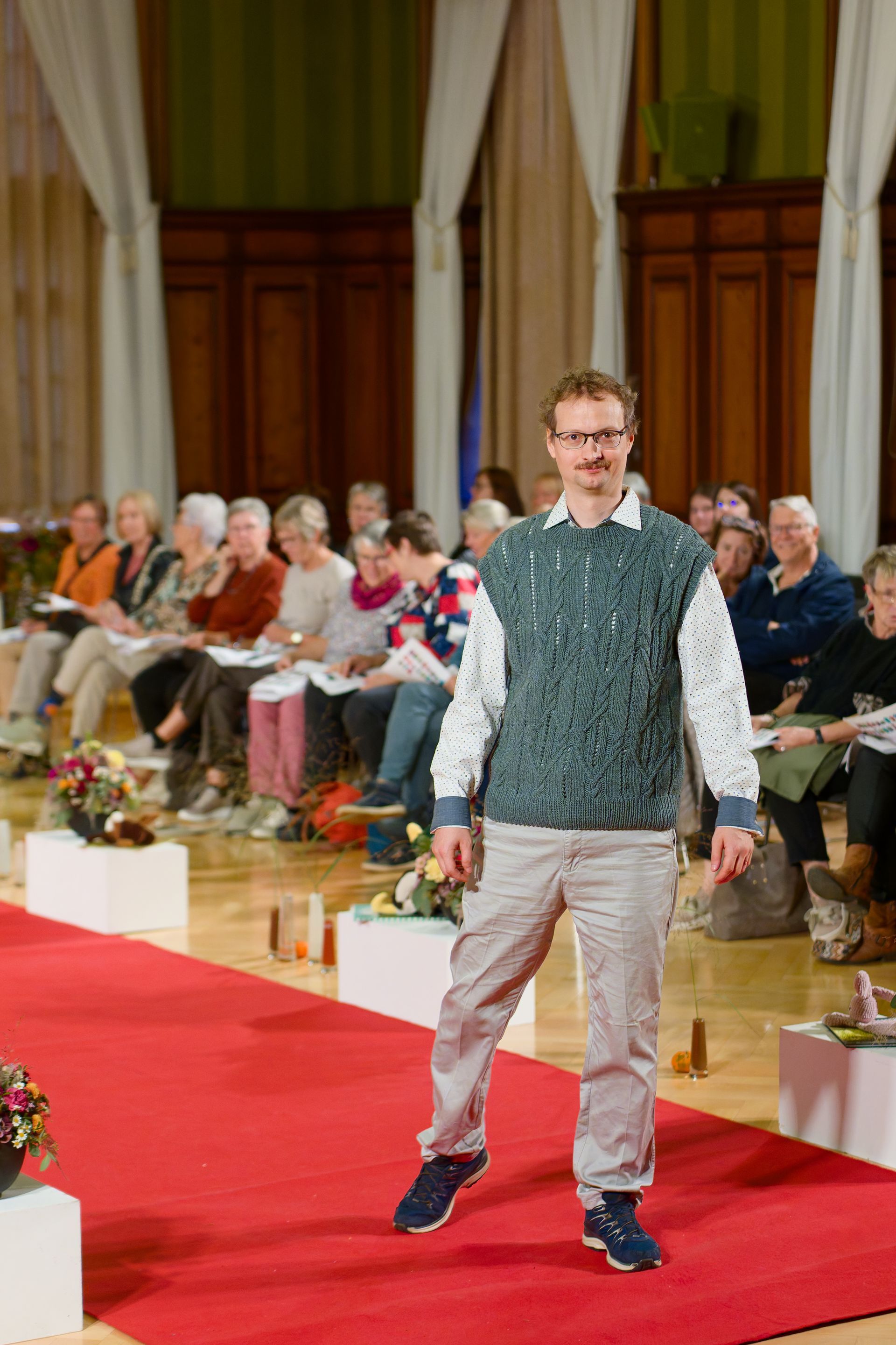 Ein Mann in grüner Strickweste, heller Hose und Brille schreitet über einen roten Teppich in einer Veranstaltungshalle.