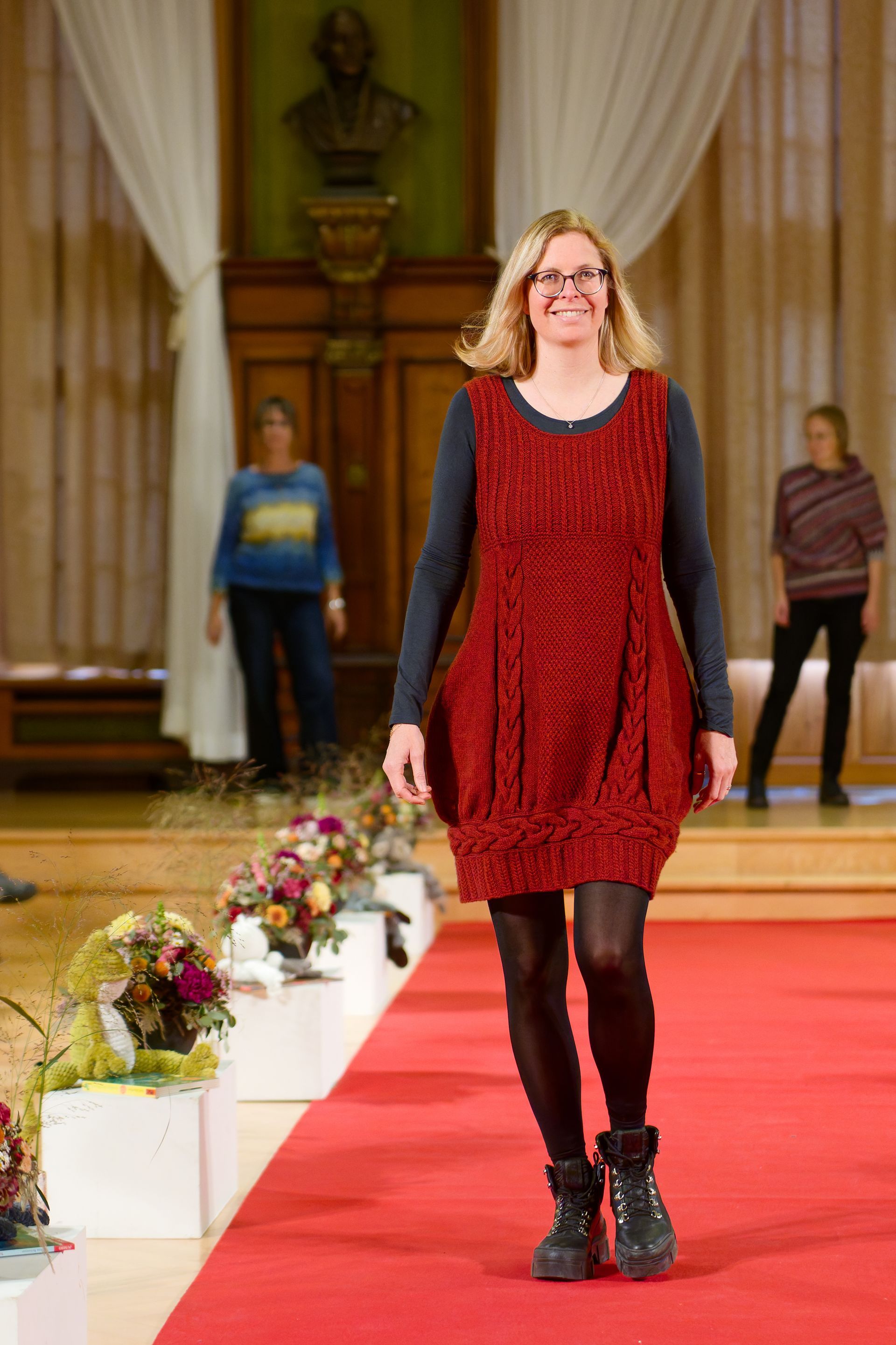 Eine Frau in einem roten, strukturierten Kleid schreitet über den roten Teppich; sie trägt schwarze Stiefel und Strumpfhosen.