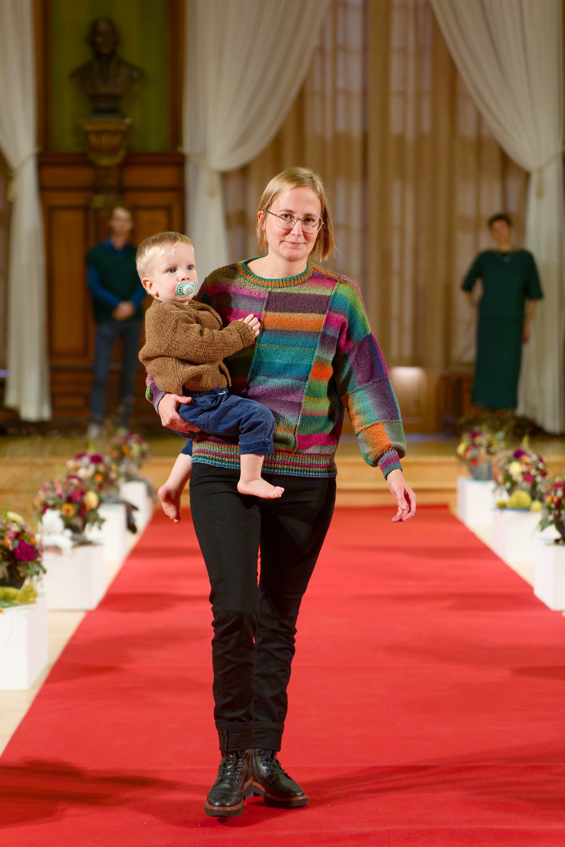 Frau in buntem Pullover trägt Baby auf dem roten Teppich.