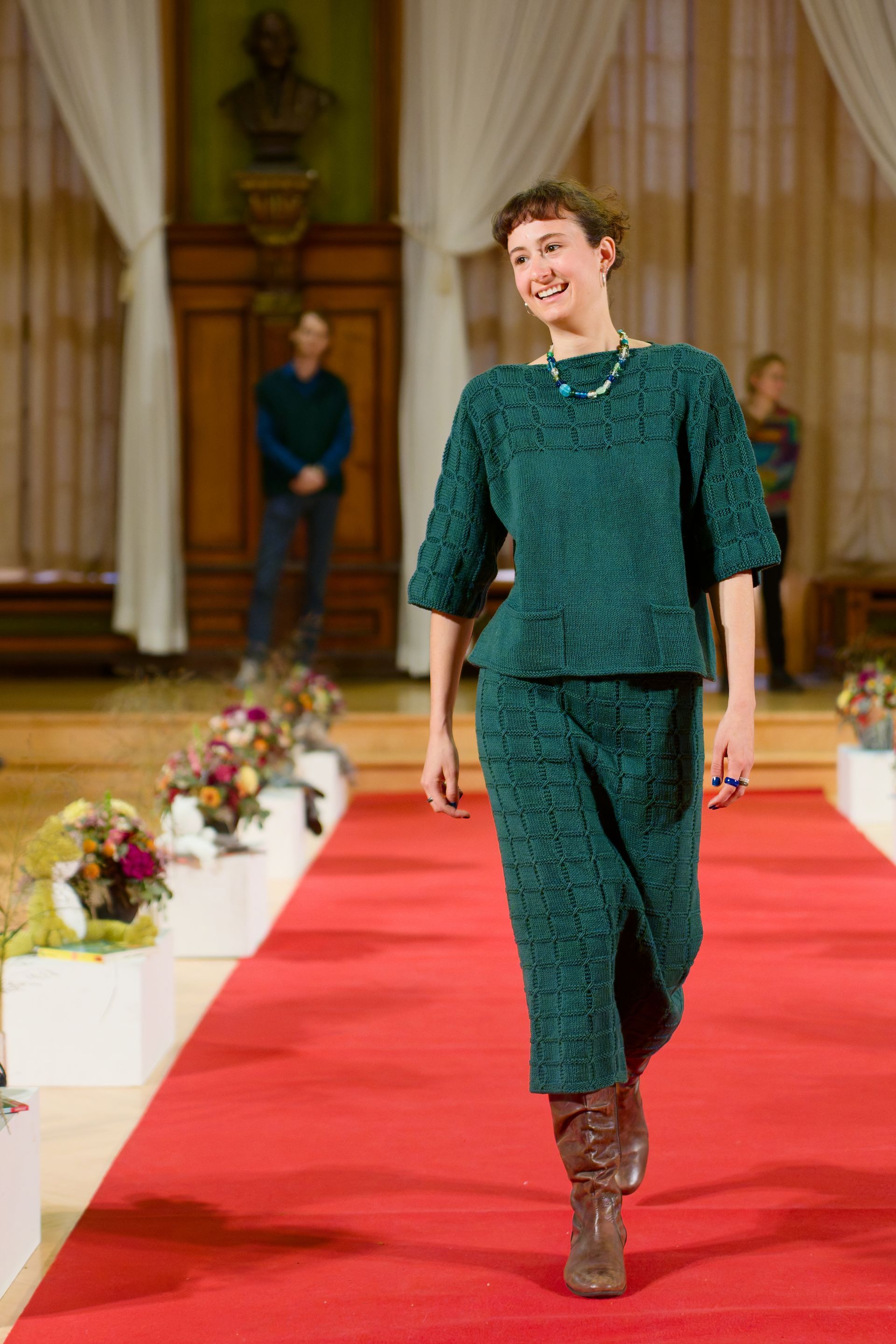 Frau in grünem Strickoutfit schreitet über einen Laufsteg; roter Teppich, Blumenarrangements.
