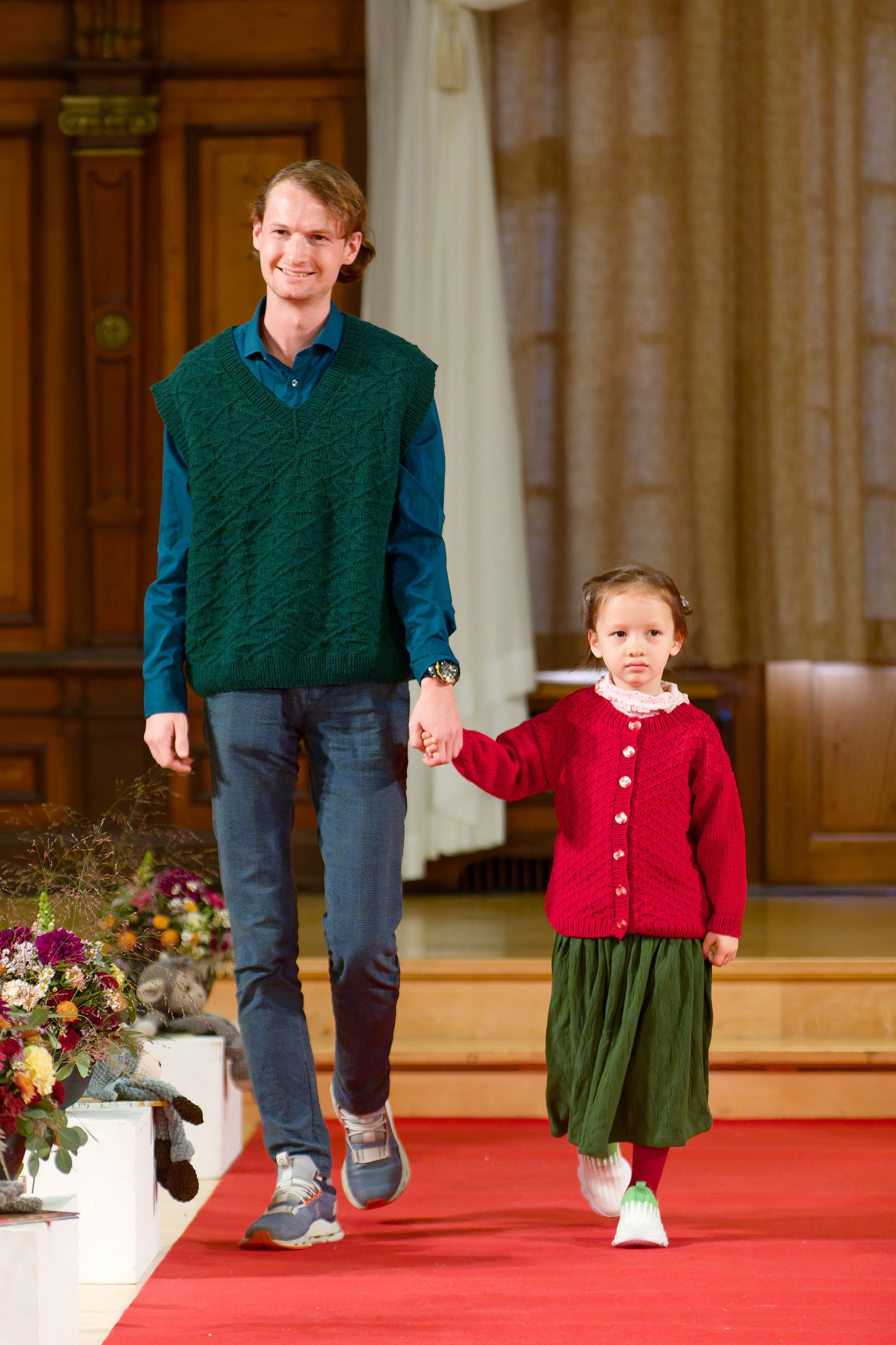 Mann und Kind gehen Hand in Hand über einen roten Teppich. Der Mann trägt eine grüne Weste, das Kind eine rote Strickjacke.